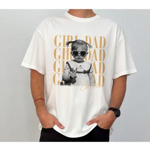 May include: White t-shirt featuring a black and white image of a child wearing heart-shaped sunglasses. The shirt has the words "GIRL DAD" in gold lettering repeated vertically, with "Est. 2020" below the image.