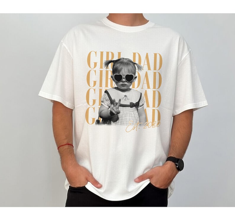 May include: White t-shirt featuring a black and white image of a child wearing heart-shaped sunglasses. The shirt has the words "GIRL DAD" in gold lettering repeated vertically, with "Est. 2020" below the image.