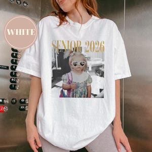 May include: White t-shirt featuring a black and white photo of a young child wearing heart-shaped sunglasses. The text "SENIOR 2026" is printed in gold above the photo. The shirt has short sleeves and a crew neck.