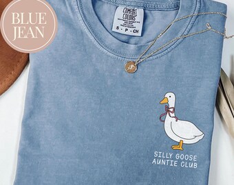 May include: A blue jean Comfort Colors t-shirt with a white goose graphic wearing a pink bow. The text "SILLY GOOSE AUNTIE CLUB" is below the goose. A gold necklace with a pendant is on the shirt.