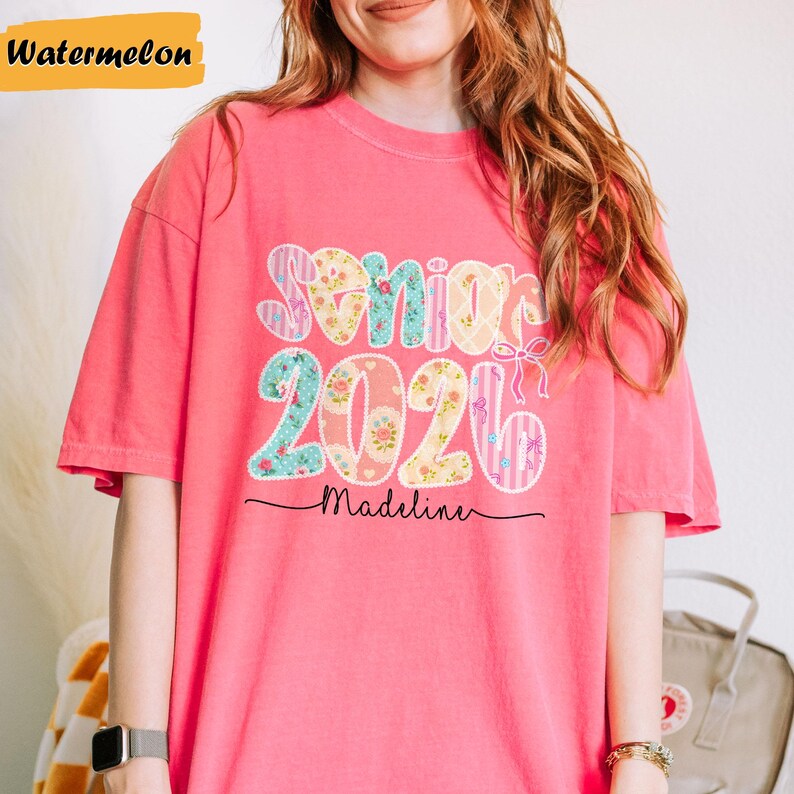 May include: A coral pink t-shirt with the words "Senior 2026" in a floral and patterned design. The name "Madeline" is written in cursive below the numbers. The word "Watermelon" is in a yellow rectangle in the top left corner.