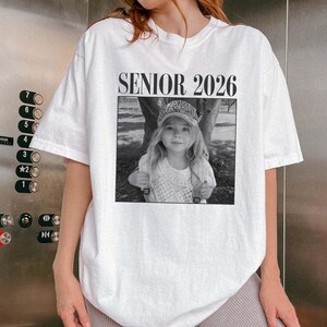 May include: White t-shirt featuring the text "SENIOR 2026" above a black and white photograph of a young girl wearing a cap. The image is centrally placed. The shirt has short sleeves and a loose fit.