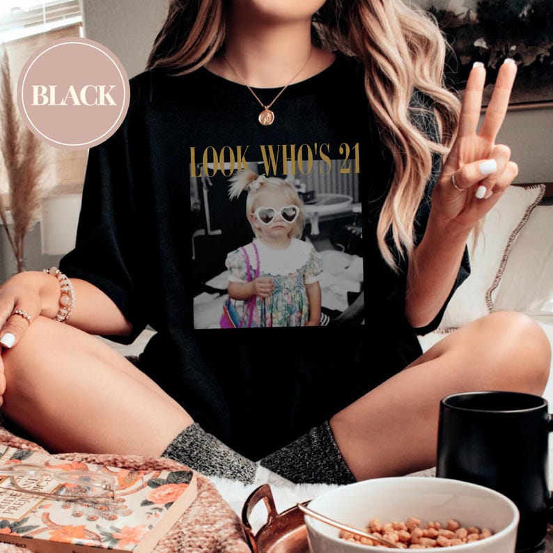 May include: Black short-sleeved t-shirt with a graphic of a young girl wearing heart-shaped sunglasses. The text "LOOK WHO'S 21" is printed in gold above the image. A round label in the upper left corner reads "BLACK".