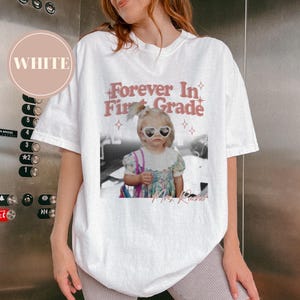 K&ouml;nnte beinhalten: Wei&szlig;es T-Shirt mit einem Aufdruck, der ein Schwarz-Wei&szlig;-Foto eines Kindes mit herzf&ouml;rmiger Sonnenbrille zeigt. Der Text lautet "Forever In First Grade" in Rosa, darunter der Name "Mrs. Rachel".