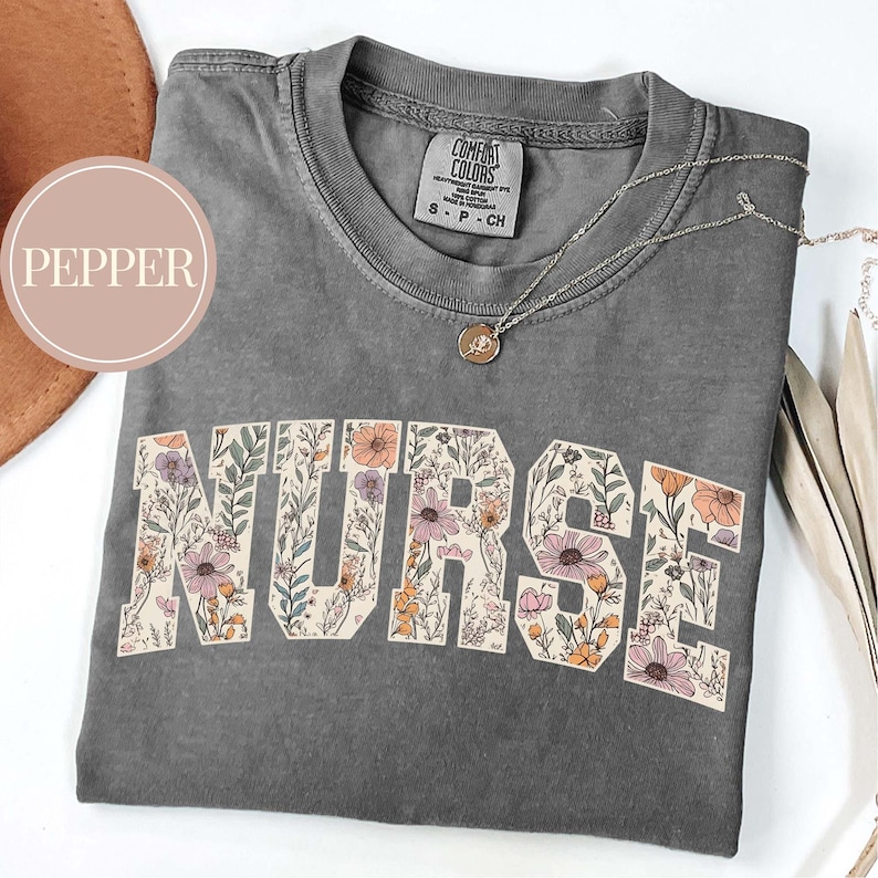 May include: A gray Comfort Colors t-shirt with the word "NURSE" in a floral design. The shirt has a crew neck and short sleeves. A silver necklace with a pendant is on the shirt.