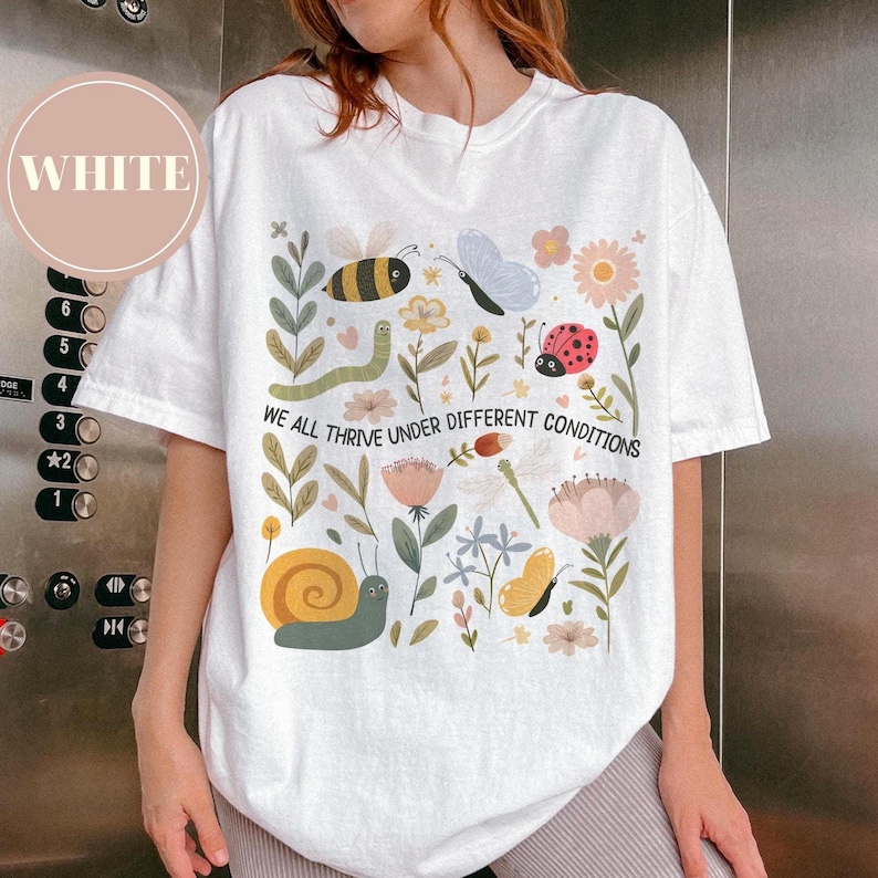 May include: White t-shirt featuring a vibrant design of flowers, insects, and the text "WE ALL THRIVE UNDER DIFFERENT CONDITIONS". The graphic includes a bee, butterfly, and ladybug.