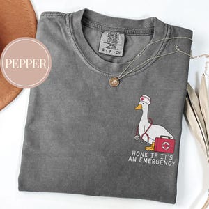 May include: A gray t-shirt with a cartoon goose wearing a nurse's hat and holding a stethoscope and a first aid kit. The text "HONK IF IT'S AN EMERGENCY" is printed below the goose.