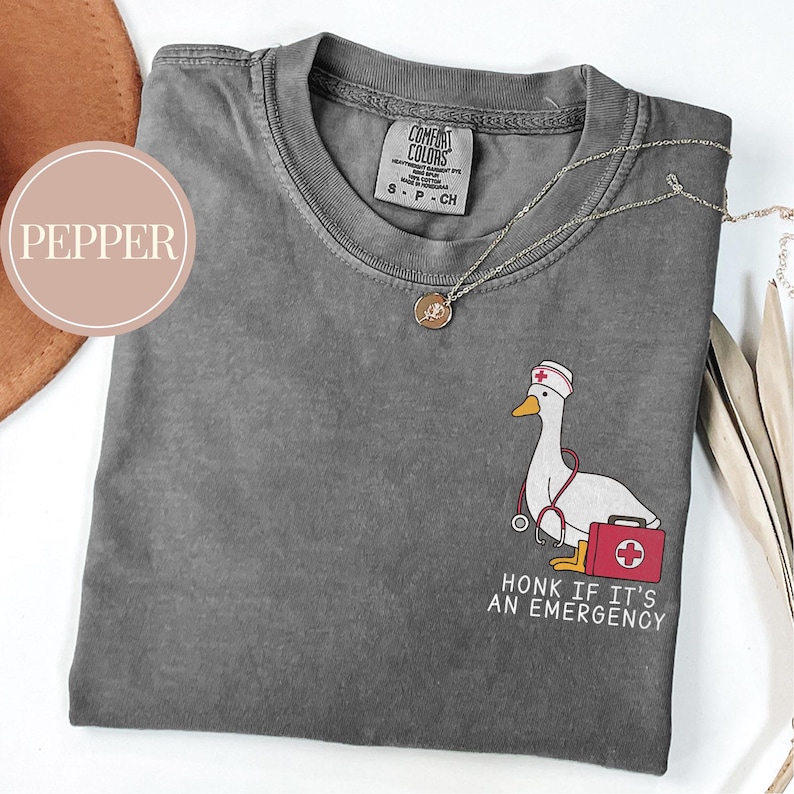 May include: A gray t-shirt with a cartoon goose wearing a nurse's hat and holding a stethoscope and a first aid kit. The text "HONK IF IT'S AN EMERGENCY" is printed below the goose.