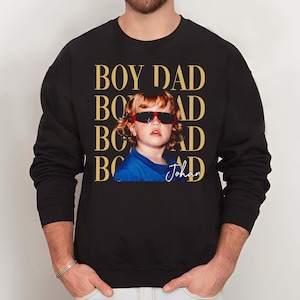 Pode incluir: Camisola preta com uma foto de uma crian&ccedil;a usando &oacute;culos de sol. As palavras "BOY DAD" s&atilde;o impressas em dourado acima da foto. O nome da crian&ccedil;a, "Johan", est&aacute; escrito abaixo. A camisola tem mangas compridas.