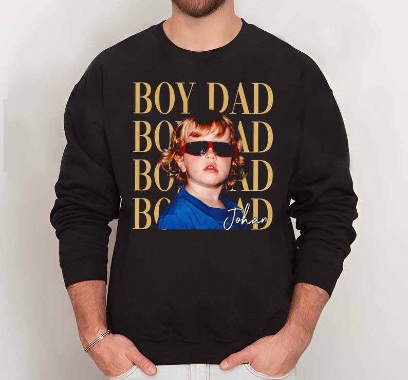 Pode incluir: Camisola preta com uma foto de uma crian&ccedil;a usando &oacute;culos de sol. As palavras "BOY DAD" s&atilde;o impressas em dourado acima da foto. O nome da crian&ccedil;a, "Johan", est&aacute; escrito abaixo. A camisola tem mangas compridas.