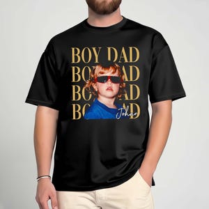 Pode incluir: T-shirt preta com a frase "BOY DAD" em letras douradas. A camisa apresenta uma foto de uma crian&ccedil;a usando &oacute;culos de sol. O nome da crian&ccedil;a, "Johan", est&aacute; escrito em it&aacute;lico abaixo.