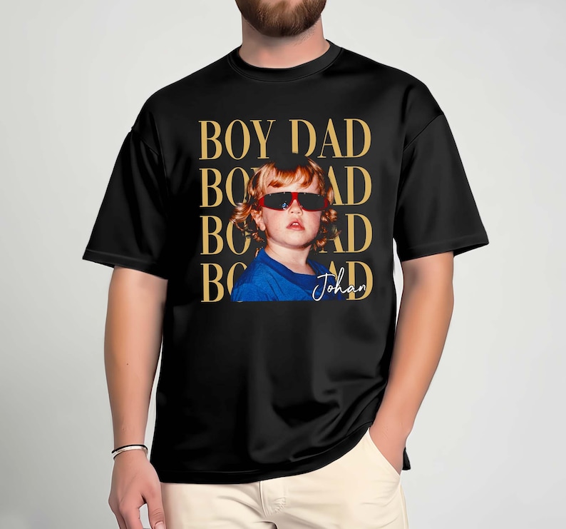 Pode incluir: T-shirt preta com a frase "BOY DAD" em letras douradas. A camisa apresenta uma foto de uma crian&ccedil;a usando &oacute;culos de sol. O nome da crian&ccedil;a, "Johan", est&aacute; escrito em it&aacute;lico abaixo.