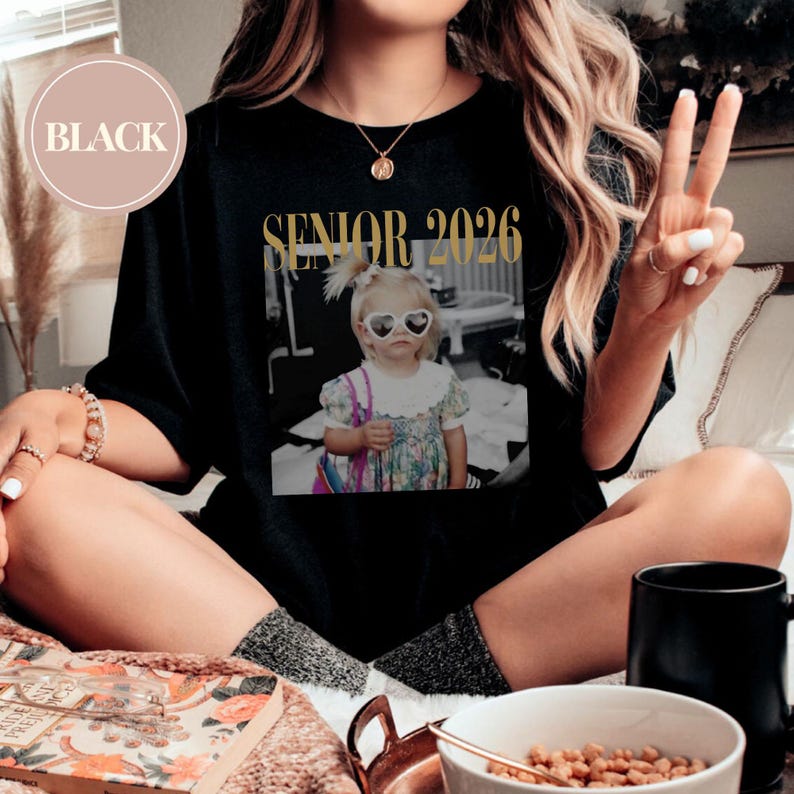 May include: Black t-shirt with a gold "SENIOR 2026" graphic and a black and white photo of a child wearing heart-shaped sunglasses. The shirt has a crew neck and short sleeves.