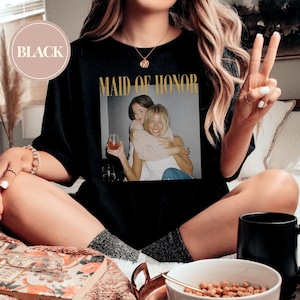 May include: Black t-shirt with the words "MAID OF HONOR" in gold lettering. The shirt features a photograph of two women embracing. A circular label in the upper left corner reads "BLACK".