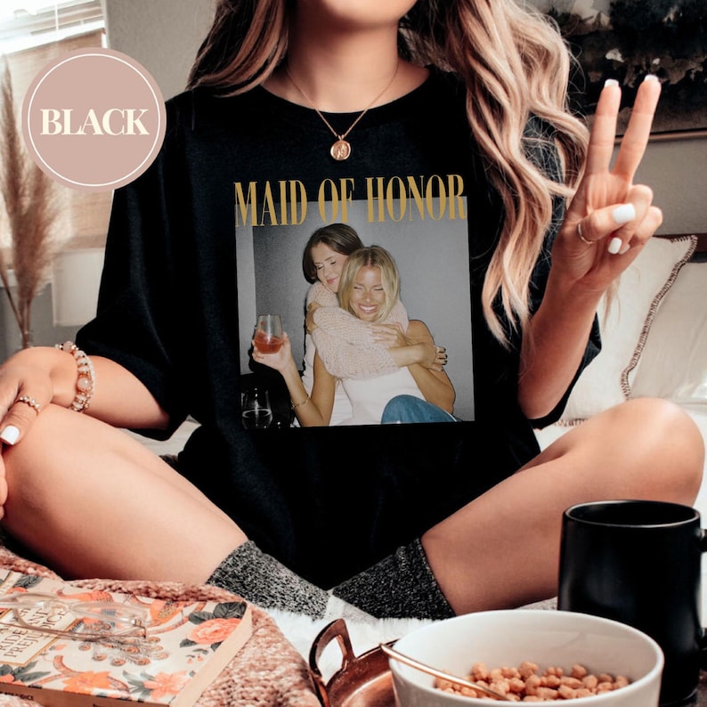 May include: Black t-shirt with the words "MAID OF HONOR" in gold lettering. The shirt features a photograph of two women embracing. A circular label in the upper left corner reads "BLACK".