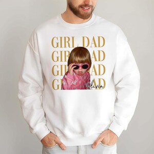 Pode incluir: Camisola branca com uma foto de uma crian&ccedil;a a usar &oacute;culos de sol cor-de-rosa e a mostrar a l&iacute;ngua. As palavras "GIRL DAD" est&atilde;o impressas a dourado, juntamente com o nome "Olivia".