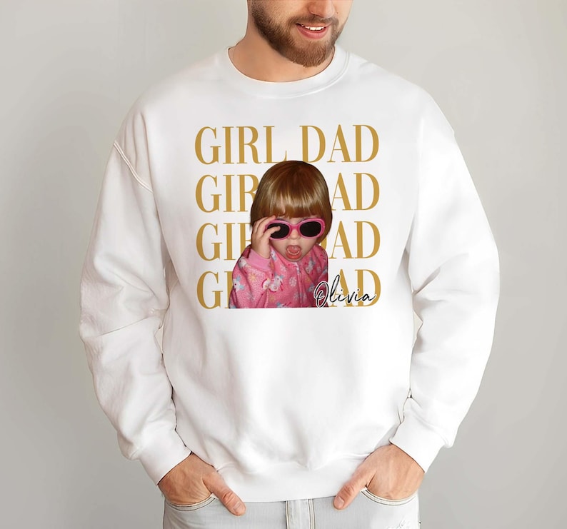 Pode incluir: Camisola branca com uma foto de uma crian&ccedil;a a usar &oacute;culos de sol cor-de-rosa e a mostrar a l&iacute;ngua. As palavras "GIRL DAD" est&atilde;o impressas a dourado, juntamente com o nome "Olivia".