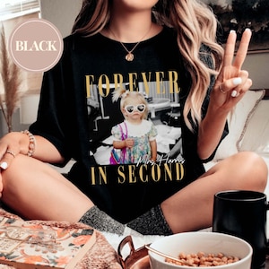 K&ouml;nnte beinhalten: Schwarzes T-Shirt mit goldfarbenem Aufdruck "FOREVER IN SECOND" &uuml;ber einem Schwarz-Wei&szlig;-Foto eines kleinen M&auml;dchens mit herzf&ouml;rmiger Sonnenbrille. Der Text "Mrs. Harris" steht unter dem Foto.