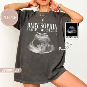 May include: Dark gray t-shirt with the text "BABY SOPHIA ARRIVING AUGUST 2025" above an ultrasound image. A small box with the words "UPLOAD PHOTO" is on the right. The shirt has a relaxed fit.