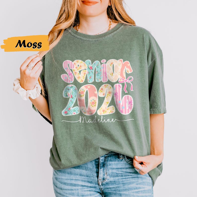 May include: Moss green t-shirt with the text "Senior 2026" in a floral and striped design. The name "Madeline" is printed below the numbers. The shirt is a comfortable, casual style.