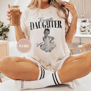 May include: Ivory-colored t-shirt with the text "Favorite Daughter" above a photo of a young girl in a tutu. The girl's name, "Emma," is written below the photo. The shirt is worn with white socks with black stripes.