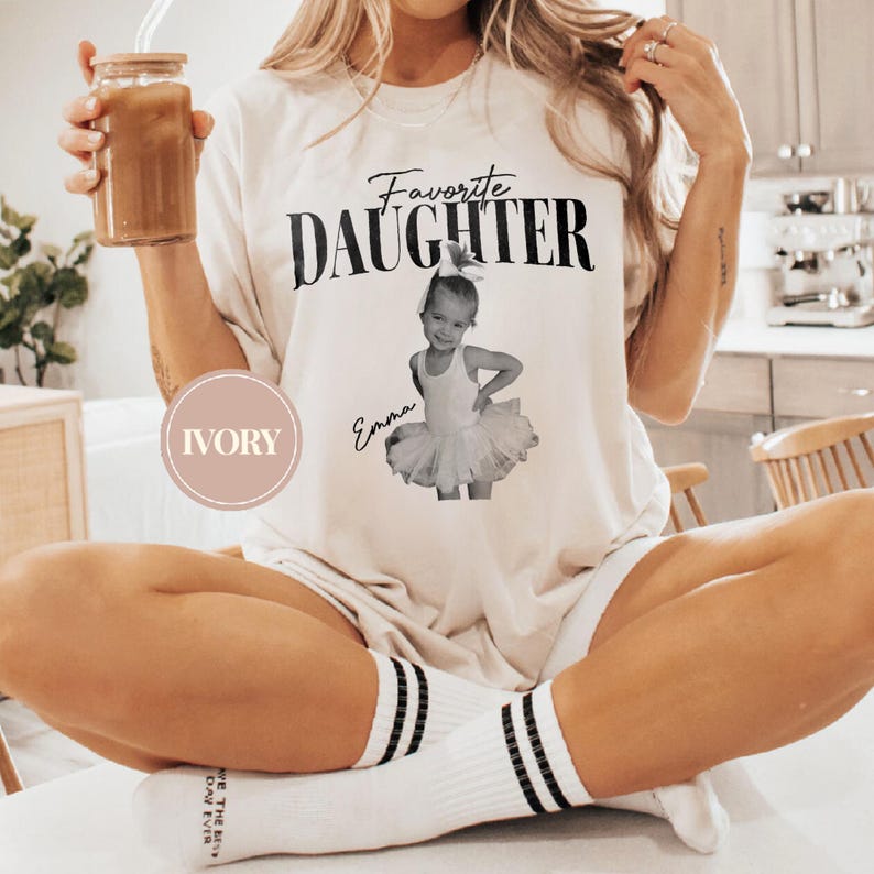 May include: Ivory-colored t-shirt with the text "Favorite Daughter" above a photo of a young girl in a tutu. The girl's name, "Emma," is written below the photo. The shirt is worn with white socks with black stripes.