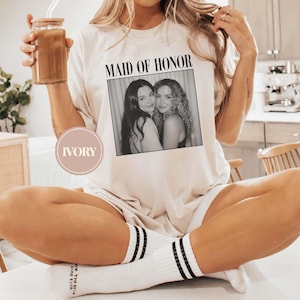 Op de afbeelding: Ivoor kleurig T-shirt met de tekst "MAID OF HONOR" boven een zwart-wit foto van twee vrouwen. Het shirt wordt gedragen met witte sokken met zwarte strepen en het woord "DAY" op de sok.