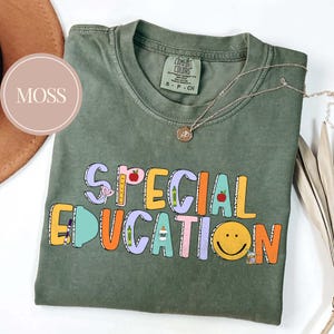 May include: Moss green t-shirt with the words "SPECIAL EDUCATION" in colorful block letters. The design includes school-themed graphics like apples, pencils, and a smiley face. A delicate silver necklace is draped across the shirt.