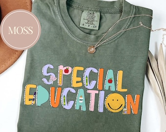 May include: Moss green t-shirt with the words "SPECIAL EDUCATION" in colorful block letters. The design includes school-themed graphics like apples, pencils, and a smiley face. A delicate silver necklace is draped across the shirt.