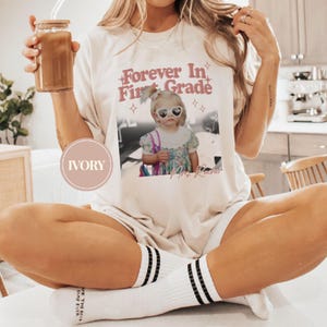 K&ouml;nnte beinhalten: Cremefarbenes T-Shirt mit dem Text "Forever In First Grade" und einem Foto eines Kindes mit herzf&ouml;rmiger Sonnenbrille. Das Shirt hat eine lockere Passform und wird mit wei&szlig;en Socken mit schwarzen Streifen getragen.
