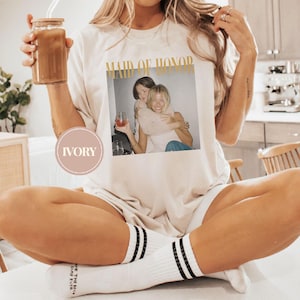 May include: Ivory-colored t-shirt with the words "MAID OF HONOR" in gold lettering. The shirt features a photo of two women embracing. The person is wearing white socks with black stripes and holding a glass jar with a brown drink.