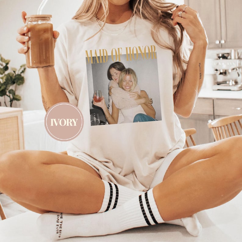 May include: Ivory-colored t-shirt with the words "MAID OF HONOR" in gold lettering. The shirt features a photo of two women embracing. The person is wearing white socks with black stripes and holding a glass jar with a brown drink.