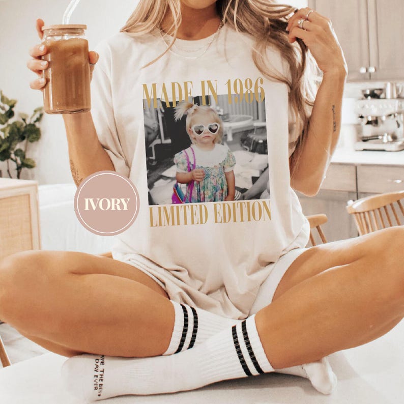 May include: Ivory-colored t-shirt with a photo print of a young child wearing heart-shaped sunglasses. The text "MADE IN 1986" and "LIMITED EDITION" are printed in gold. The person is wearing white socks with black stripes.