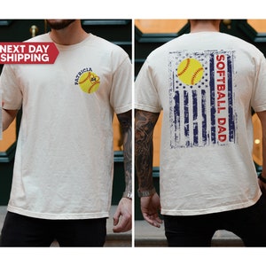May include: A white t-shirt with a softball graphic and the text "PATRICIA 24" on the front. The back of the shirt features a distressed American flag design with the text "SOFTBALL DAD".