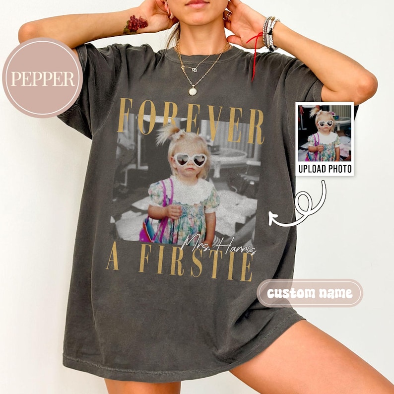 May include: Dark gray t-shirt with a vintage-style photo print. The photo features a young girl wearing heart-shaped sunglasses. Gold text reads "FOREVER A FIRSTIE" above and below the image. A small photo is on the right with "UPLOAD PHOTO" text.