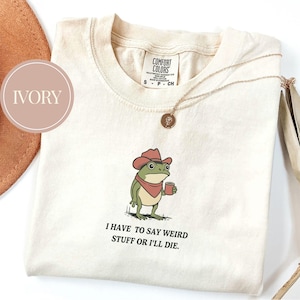 May include: Ivory-colored t-shirt featuring a cartoon frog wearing a cowboy hat and holding a mug. The frog has a red bandana and the text "I HAVE TO SAY WEIRD STUFF OR I'LL DIE." A gold necklace with a pendant is draped on the shirt.