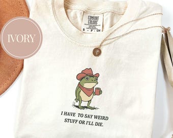 Puede incluir: Camiseta de color marfil con una rana de dibujos animados con sombrero de vaquero y taza. La rana tiene un pañuelo rojo y el texto "I HAVE TO SAY WEIRD STUFF OR I'LL DIE." Un collar dorado con colgante está sobre la camiseta.