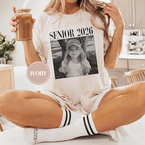 May include: Ivory t-shirt featuring the text "SENIOR 2026" above a black and white photograph of a young girl. The girl is wearing a hat and smiling. The person is wearing white socks with black stripes.