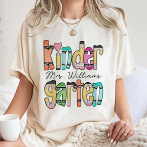 May include: A cream-colored short-sleeve t-shirt featuring a graphic print with the text "Kindergarten" and "Mrs. Williams" in the center. The letters are styled to resemble colorful crayons with a small pink heart above the word Kinder.