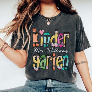 May include: A charcoal grey short-sleeve t-shirt featuring a colorful graphic design with the text "Kindergarten" styled to look like crayons, a small pink heart, and the name "Mrs. Williams" printed in white cursive script in the center.