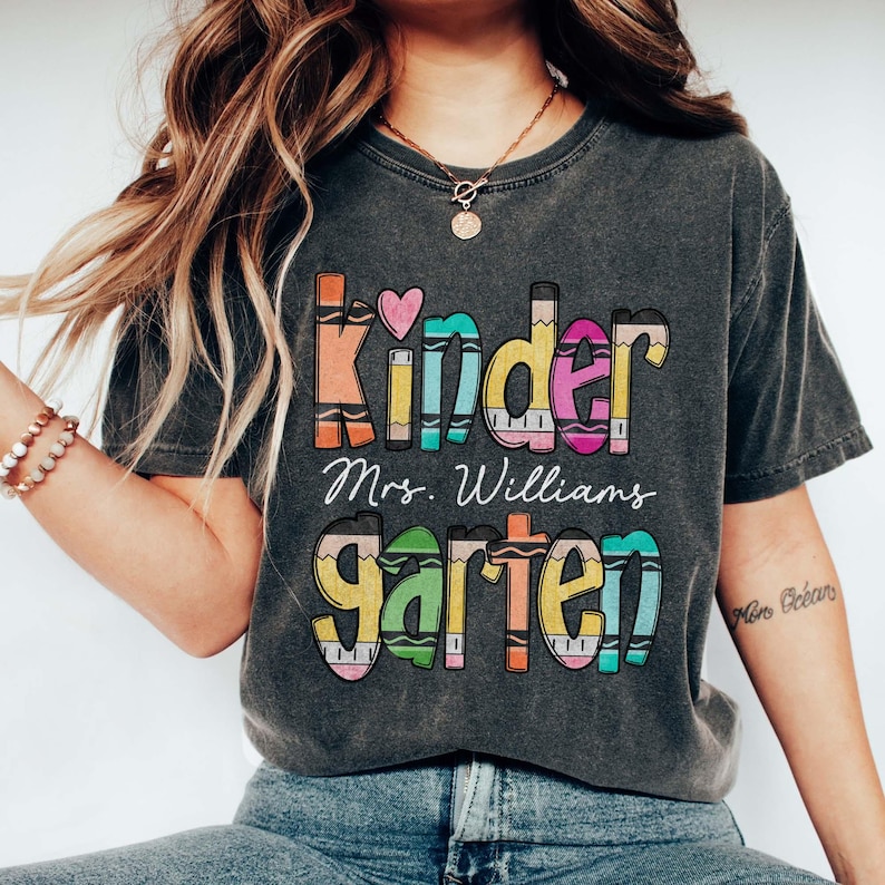 May include: A charcoal grey short-sleeve t-shirt featuring a colorful graphic design with the text "Kindergarten" styled to look like crayons, a small pink heart, and the name "Mrs. Williams" printed in white cursive script in the center.