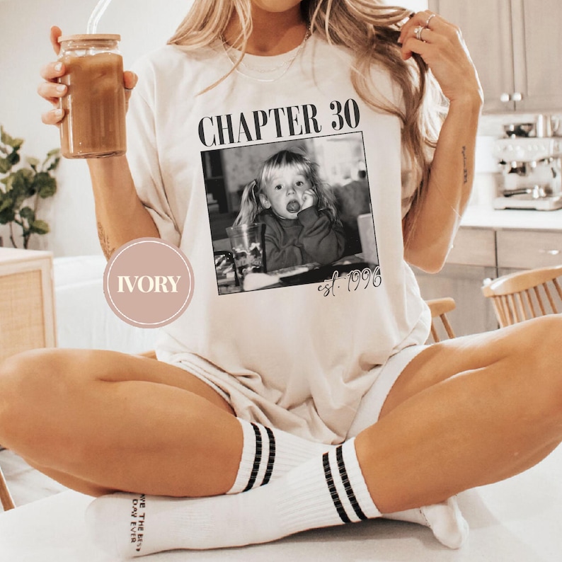 May include: Ivory-colored t-shirt with the text "CHAPTER 30" above a black and white photo of a young child. The text "est. 1993" is below the photo. The person is wearing white socks with black stripes.
