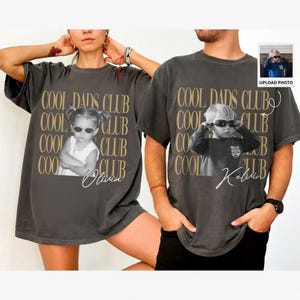 May include: Two charcoal gray t-shirts with the text "COOL DADS CLUB" in gold. Each shirt features a black and white photo of a child wearing sunglasses. One shirt has the name "Olivia" and the other has "Kelvin".