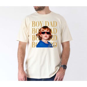 Pode incluir: Camiseta creme com um gr&aacute;fico de uma crian&ccedil;a usando &oacute;culos de sol. As palavras "BOY DAD" s&atilde;o impressas verticalmente em dourado, com o nome da crian&ccedil;a, "Johan", em it&aacute;lico. A crian&ccedil;a est&aacute; vestindo uma camisa azul.