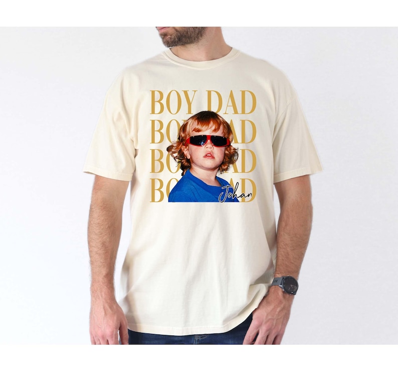 Pode incluir: Camiseta creme com um gr&aacute;fico de uma crian&ccedil;a usando &oacute;culos de sol. As palavras "BOY DAD" s&atilde;o impressas verticalmente em dourado, com o nome da crian&ccedil;a, "Johan", em it&aacute;lico. A crian&ccedil;a est&aacute; vestindo uma camisa azul.