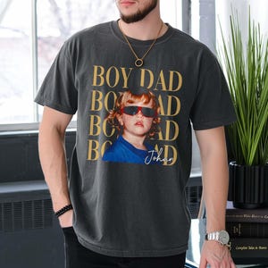 Pode incluir: T-shirt cinzenta escura com o texto "BOY DAD" dourado repetido verticalmente. Uma foto de uma crian&ccedil;a usando &oacute;culos de sol vermelhos e uma camisa azul &eacute; impressa na frente. O nome da crian&ccedil;a, "Johan", est&aacute; escrito em it&aacute;lico.