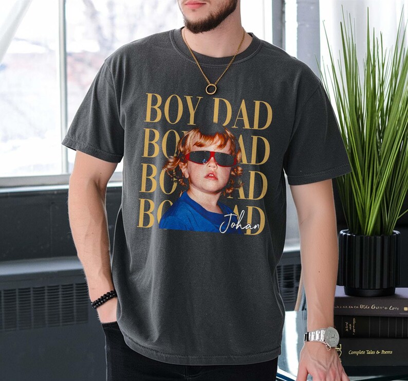 Pode incluir: T-shirt cinzenta escura com o texto "BOY DAD" dourado repetido verticalmente. Uma foto de uma crian&ccedil;a usando &oacute;culos de sol vermelhos e uma camisa azul &eacute; impressa na frente. O nome da crian&ccedil;a, "Johan", est&aacute; escrito em it&aacute;lico.