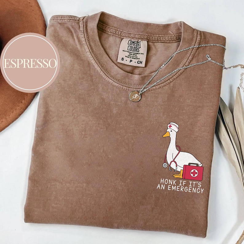 May include: A brown t-shirt with a distressed look featuring a cartoon goose wearing a nurse's hat and holding a stethoscope and a first aid kit. The text "Honk if it's an emergency" is printed below the goose.