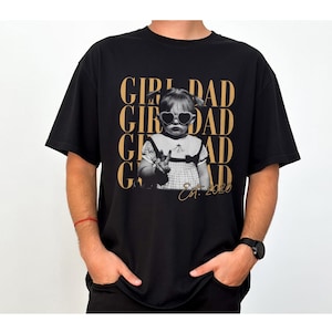 May include: Black t-shirt featuring a black and white image of a child wearing heart-shaped sunglasses, with the words "GIRL DAD" in gold lettering. The text "Est. 2020" is also included.