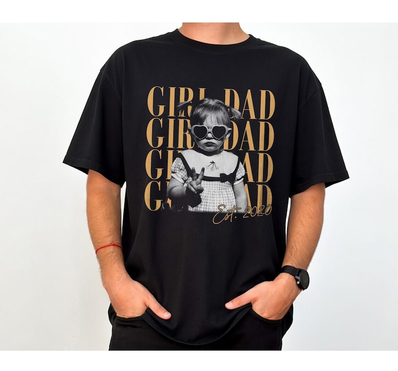 May include: Black t-shirt featuring a black and white image of a child wearing heart-shaped sunglasses, with the words "GIRL DAD" in gold lettering. The text "Est. 2020" is also included.